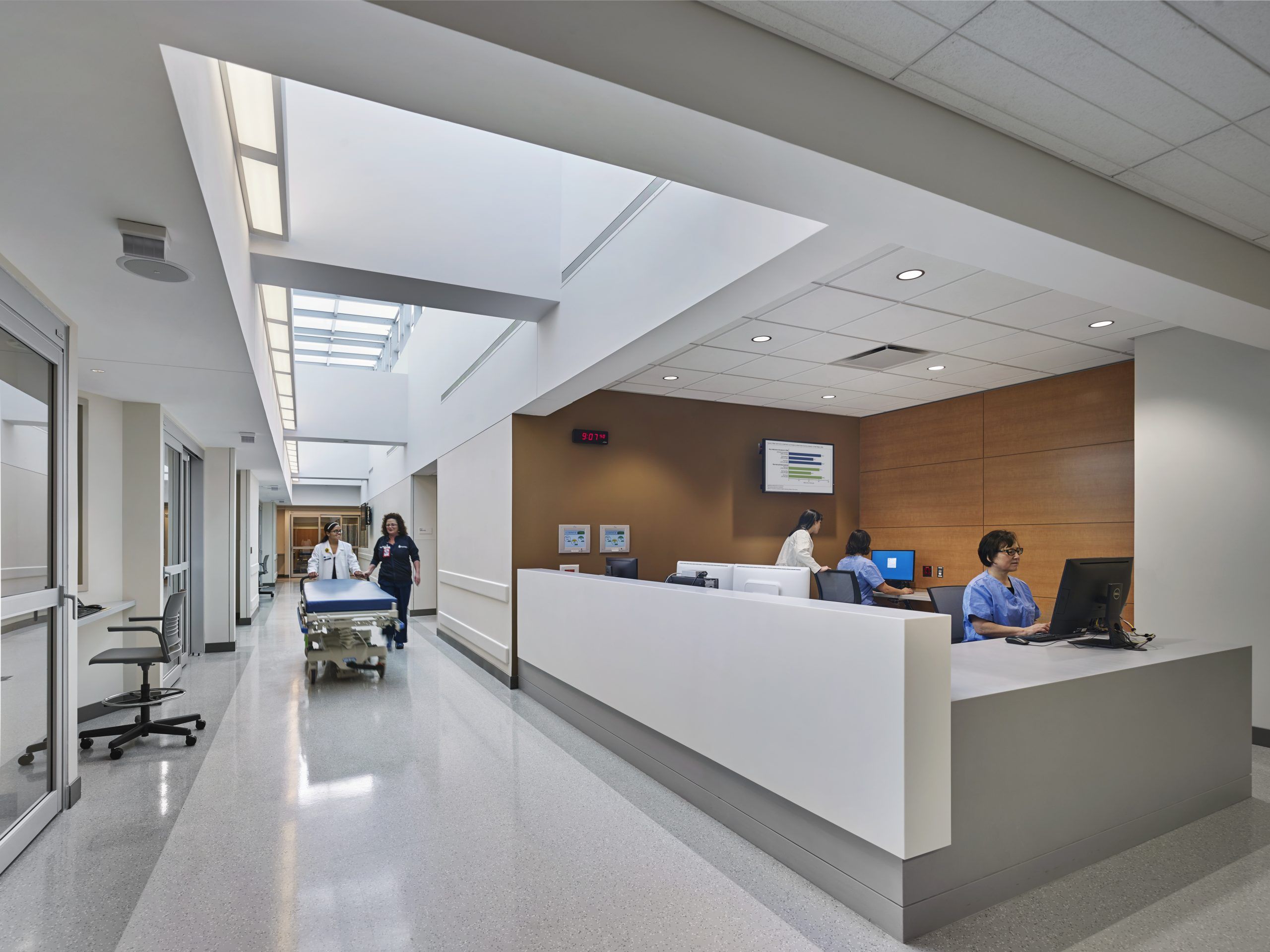 A nurse and doctor push a gurney through a hallway lit naturally by a skylight
